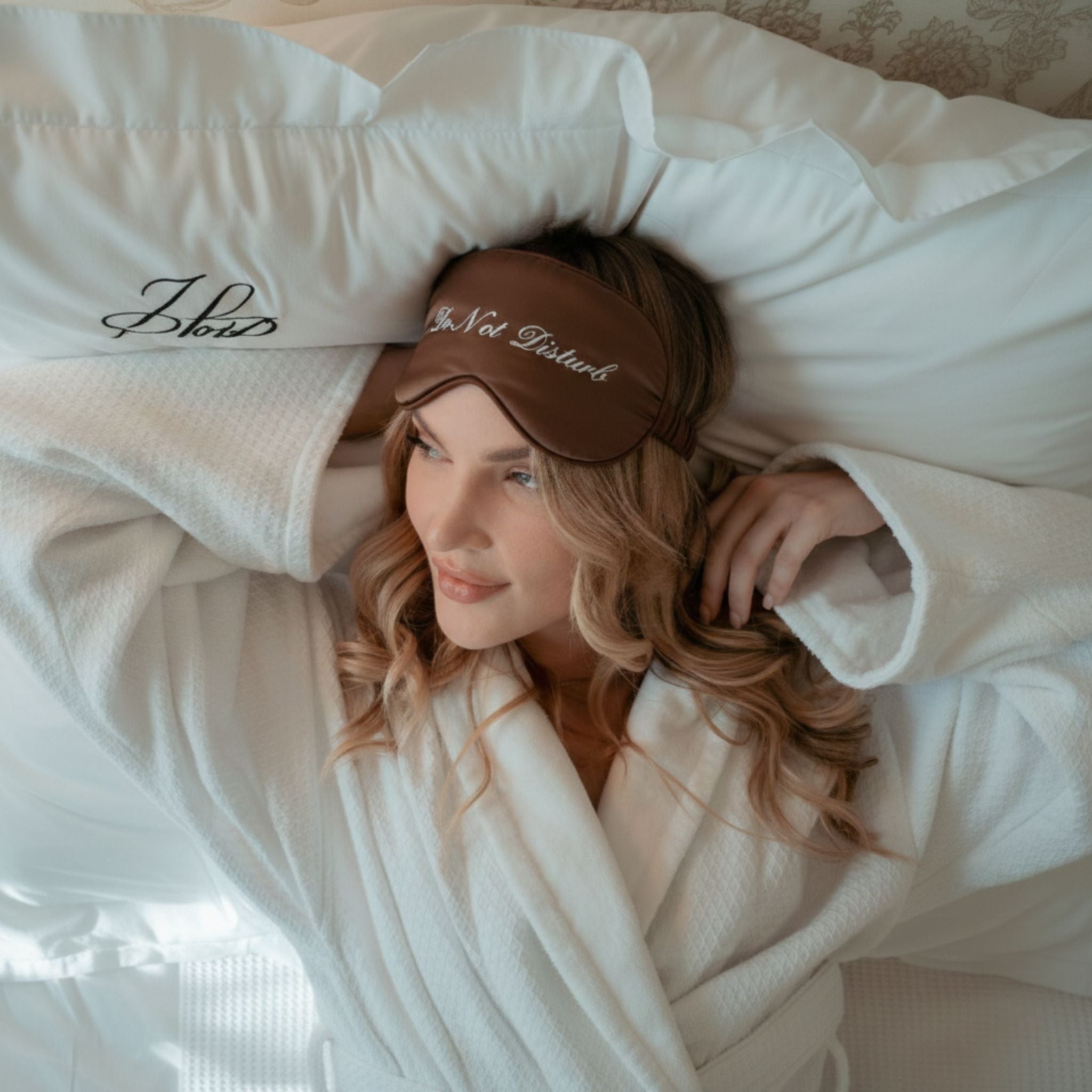 Brown silk eye mask with 'Do Not Disturb' text, worn by a woman in a cozy white robe on a plush pillow.
