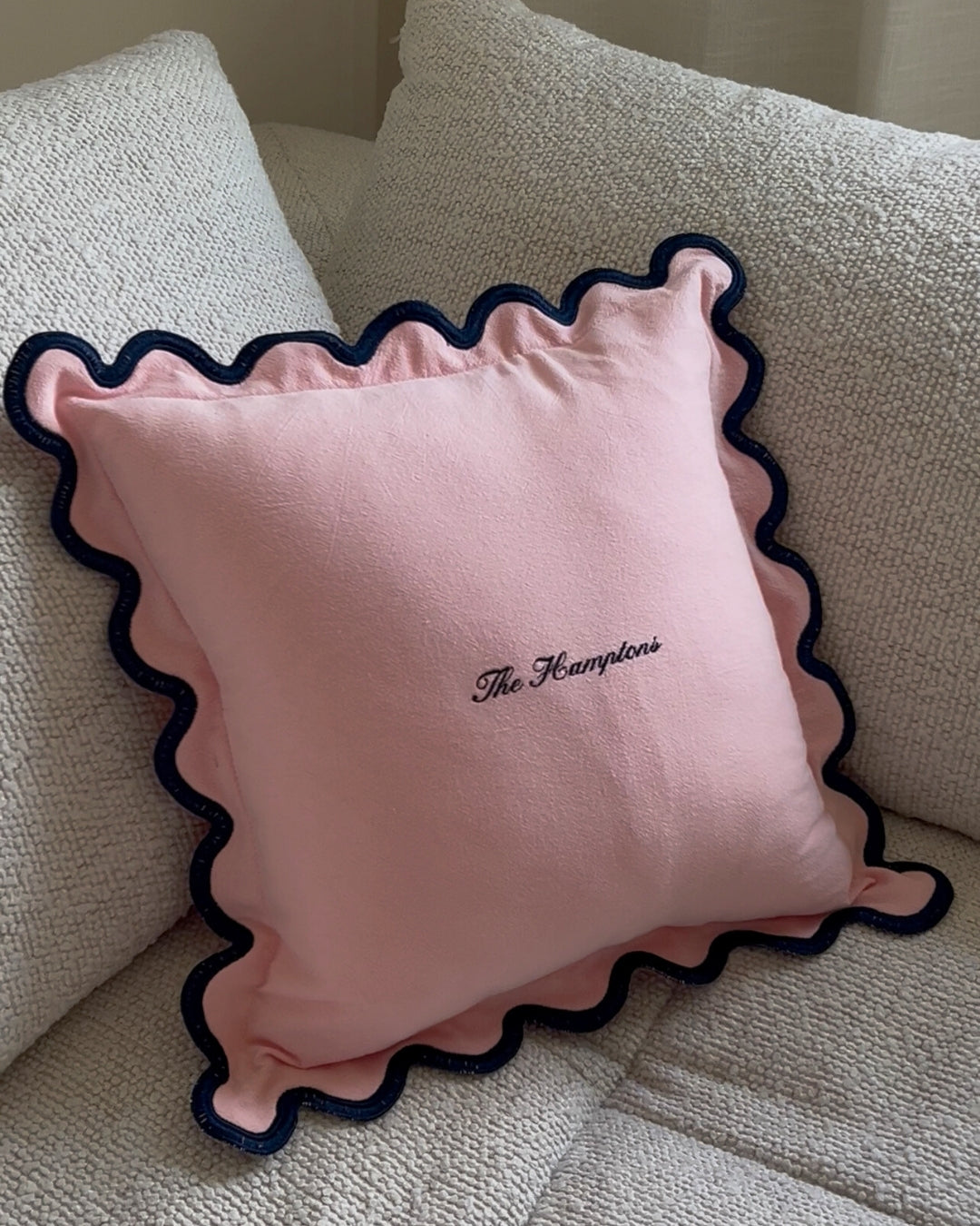 Pink decorative pillow with navy ruffled edges and embroidered 'The Hamptons' text.