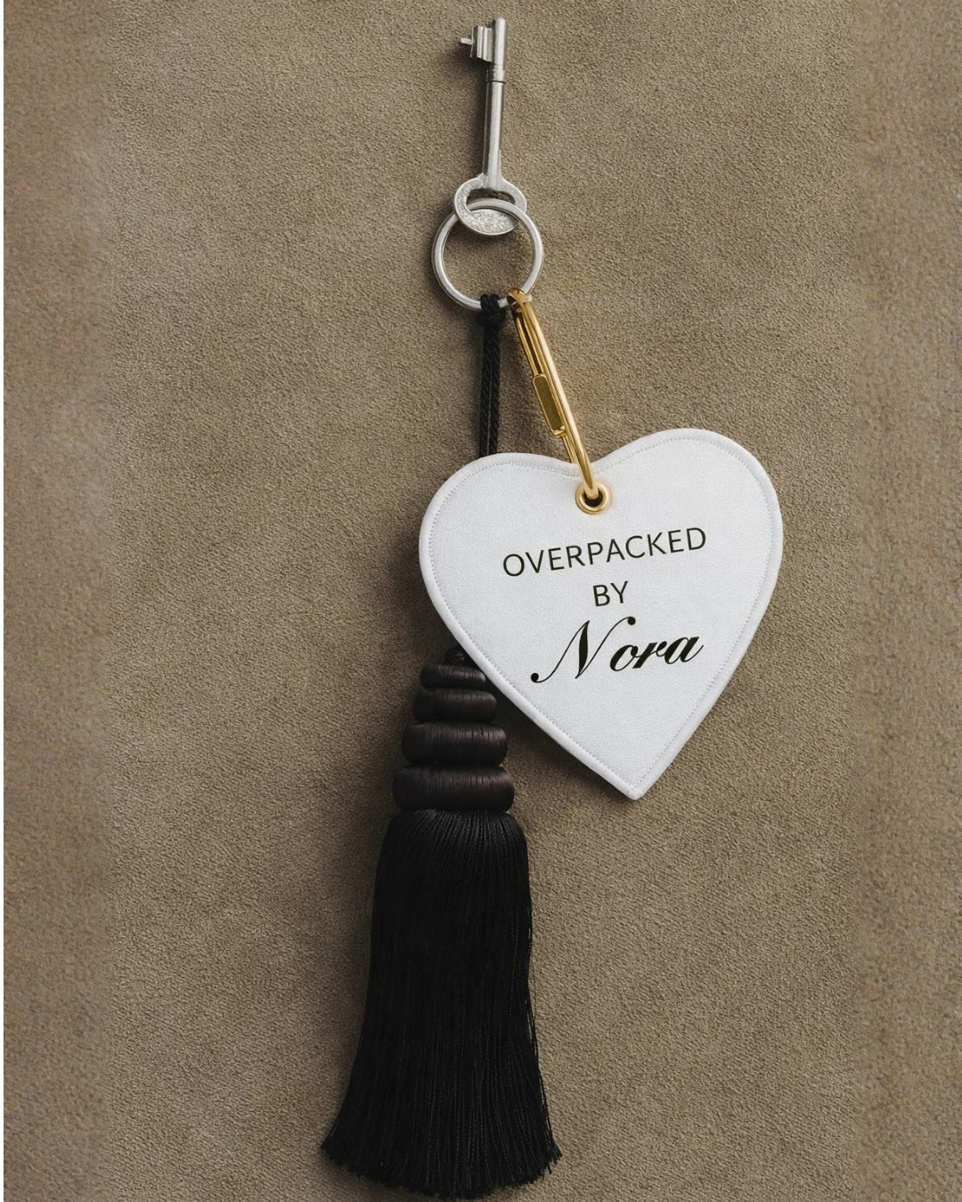 Heart-shaped keychain tag in white with black tassel, featuring 'OVERPACKED BY Nora' text.