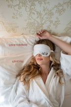 White striped silk sleep mask with monogram 'A.B.' on a woman in a cozy white robe, resting on embroidered pillows.