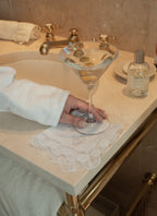 Elegant martini with olives on a lace napkin by a marble sink, featuring a cozy robe and grooming products.