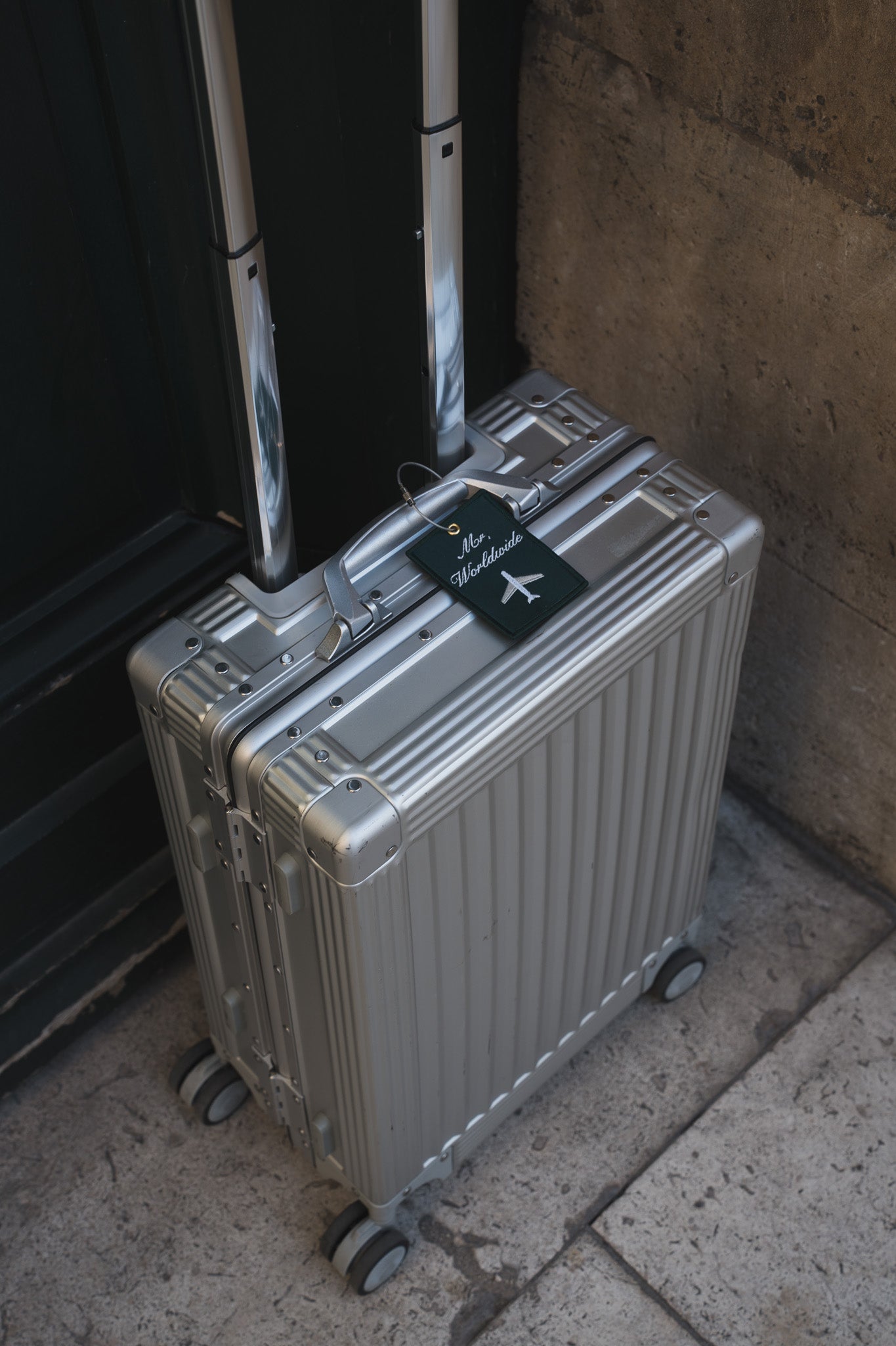 Silver hard-shell luggage with retractable handle and wheels, featuring a green travel tag.