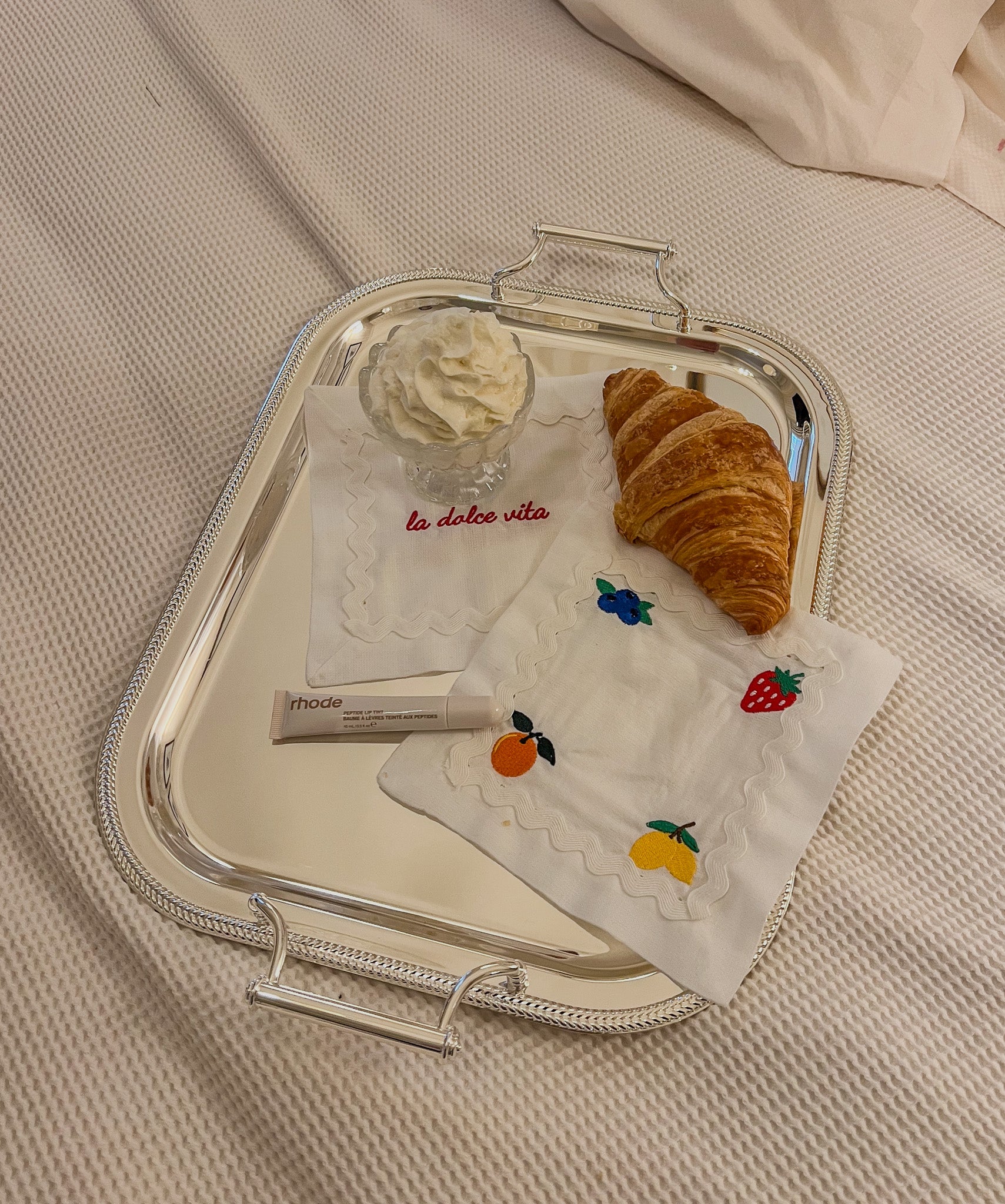 Elegant silver tray with croissant, whipped cream, and embroidered napkins featuring fruit designs.