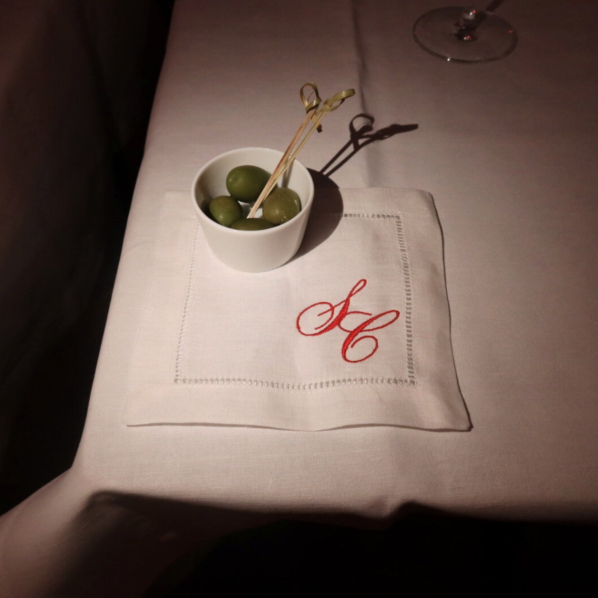White bowl with green olives on a white napkin with red monogram, on a dark surface.