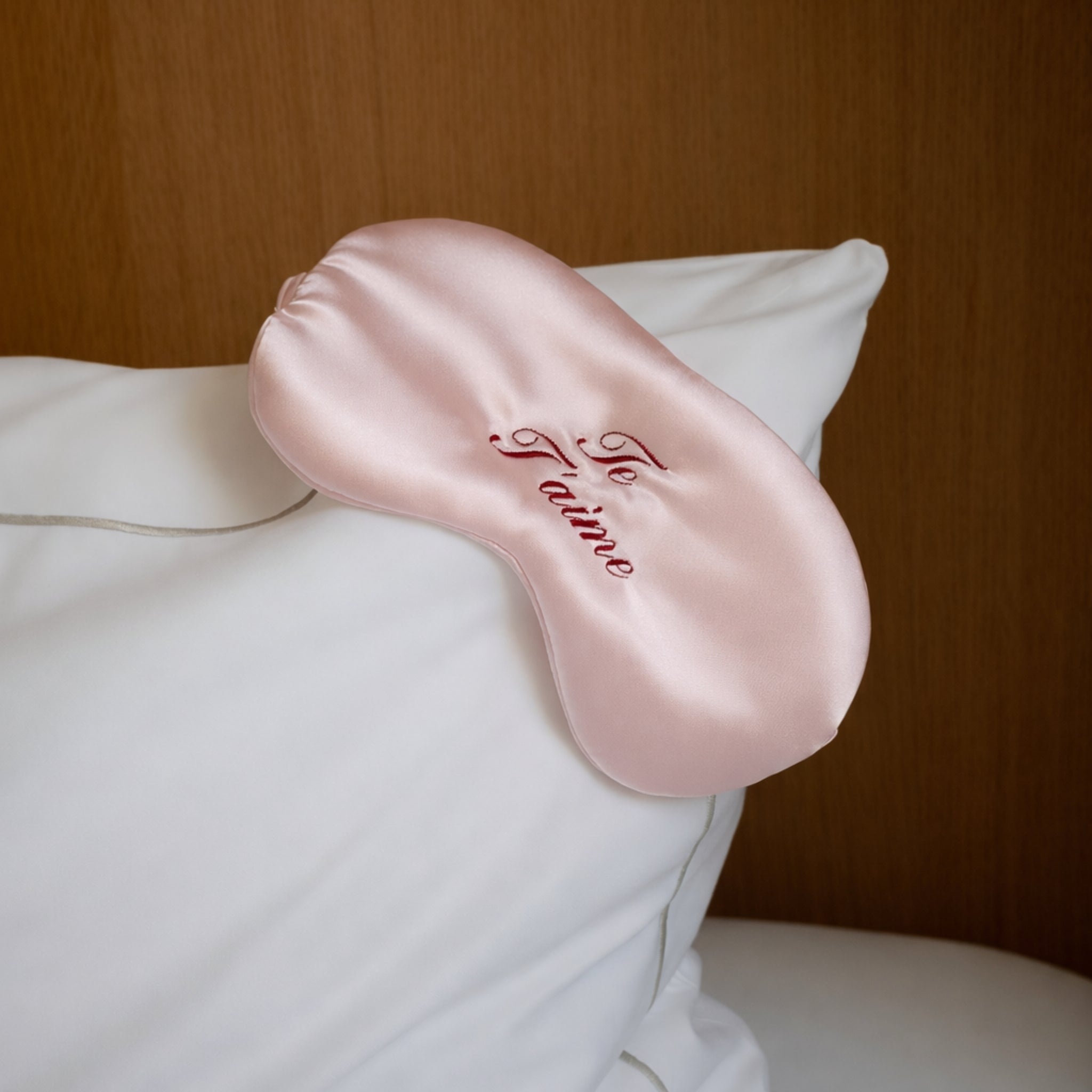 Pink satin eye mask with 'Je T'aime' embroidery on a white pillow against a wooden background