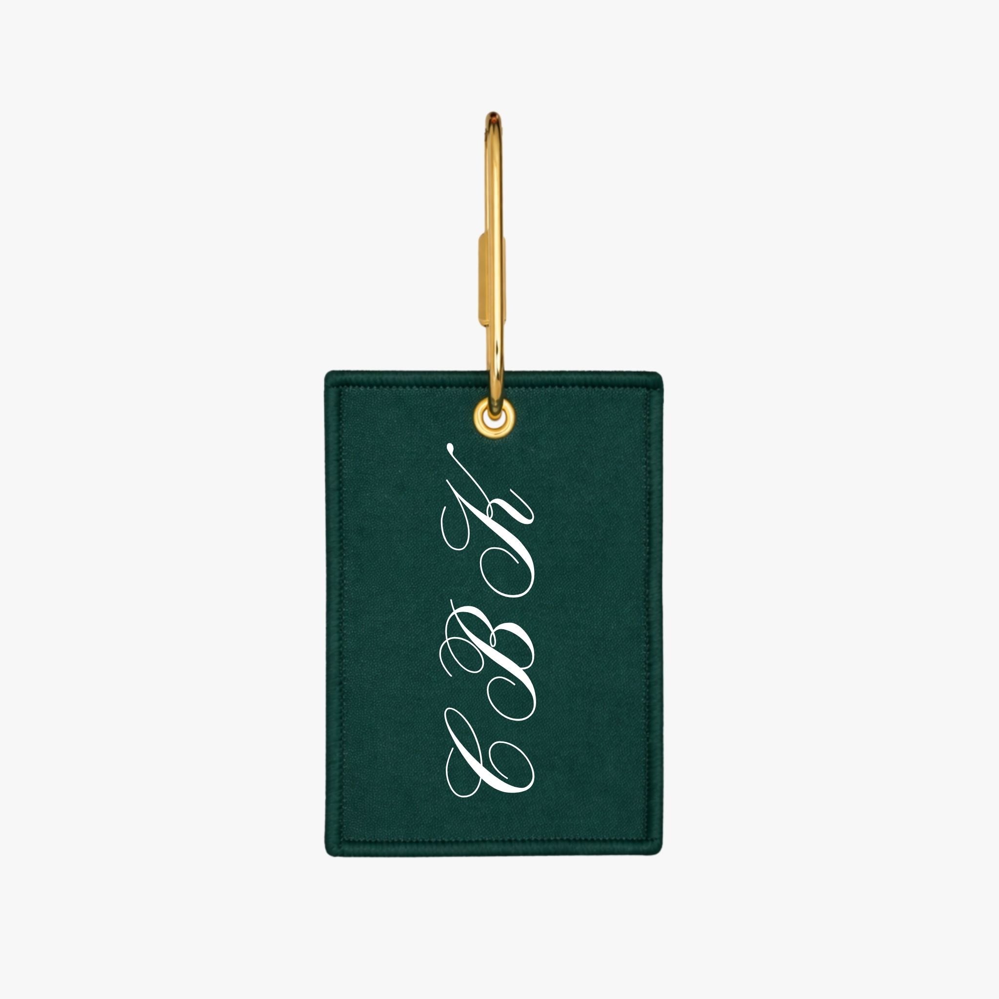 Personalized green leather luggage tag with white cursive initials and gold hardware.