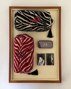 A framed display containing two zip pouches with zebra-style patterns, a small dark rectangular box with gold initials, and two small black-and-white photo cards, all arranged on a beige backing inside a wood frame.