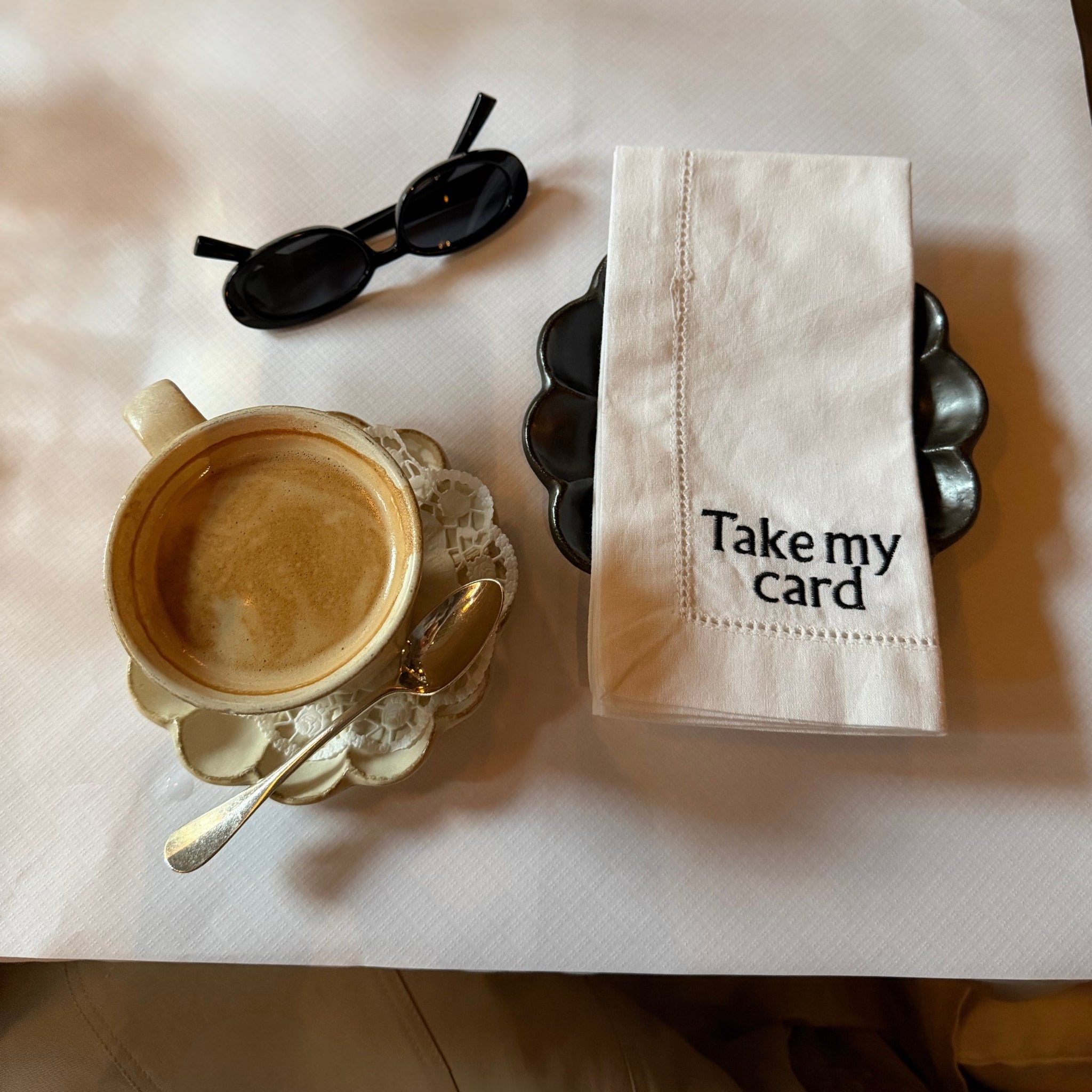Stylish coffee cup with saucer, silver spoon, and napkin reading 'Take my card' and black sunglasses.