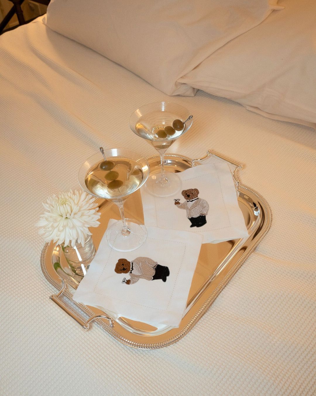 Embroidered bear cocktail napkins in white, elegant drinkware, and a flower on a silver tray.