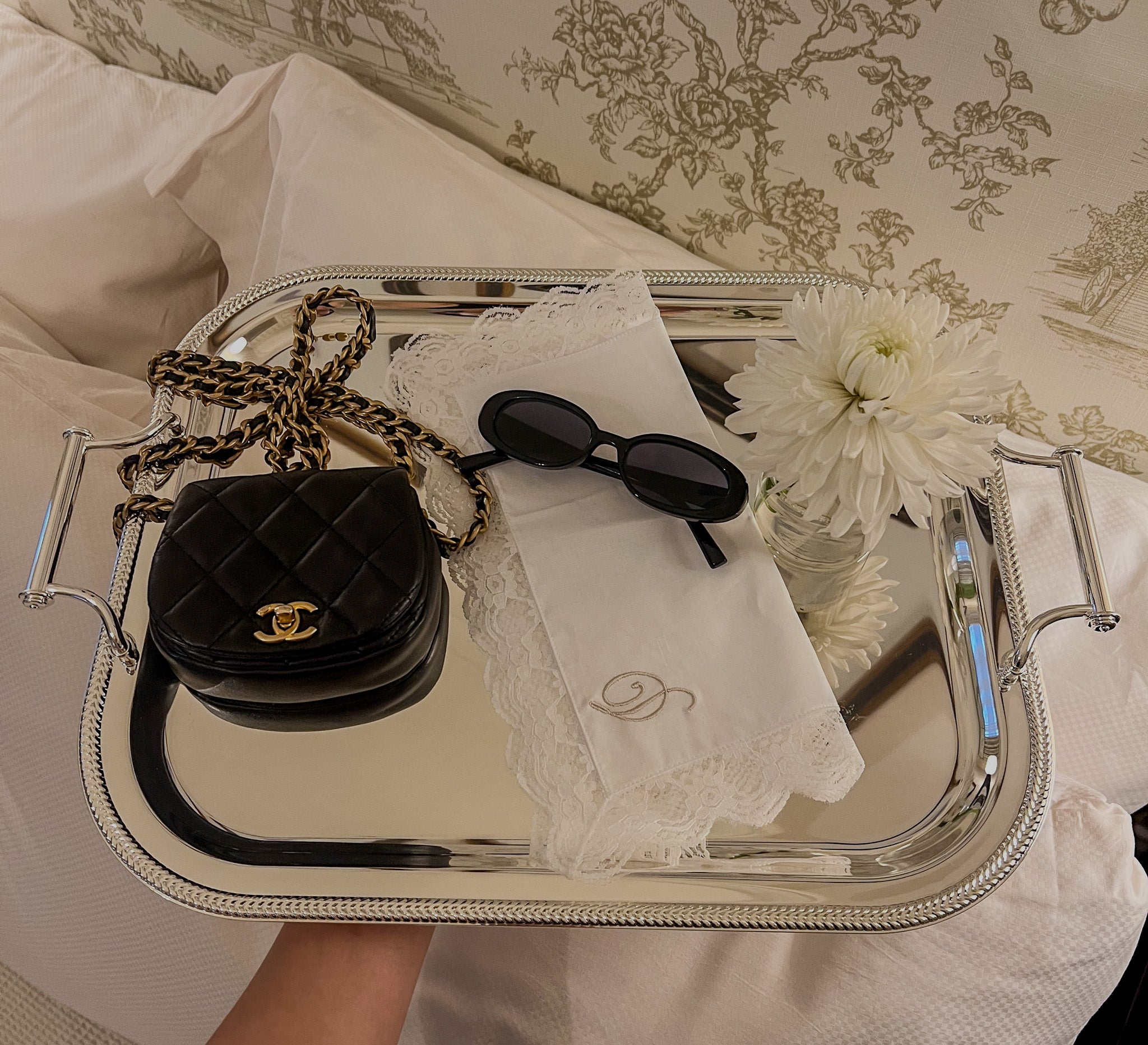 Elegant silver tray with black quilted handbag, sunglasses, lace napkin, and white flower centerpiece.