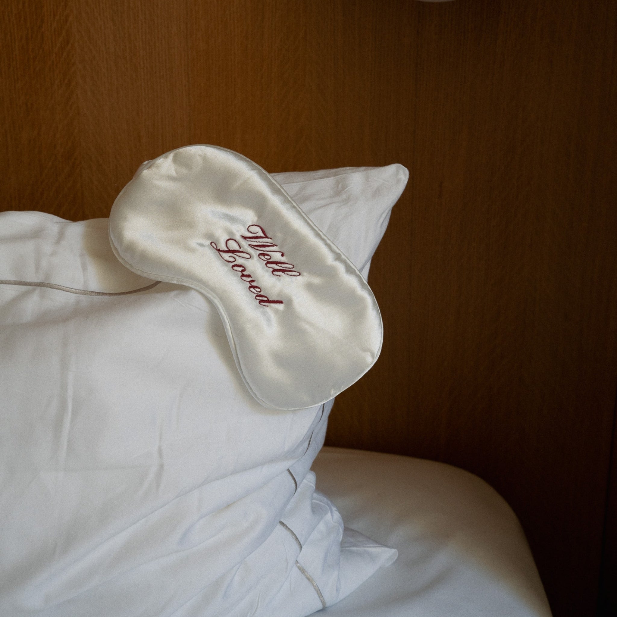 White satin eye mask with 'Most Loved' embroidery on a white pillow against a wooden background